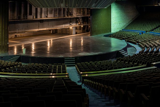 Empty theater stage with rows of seats, stage floor reflects light, dark interior.