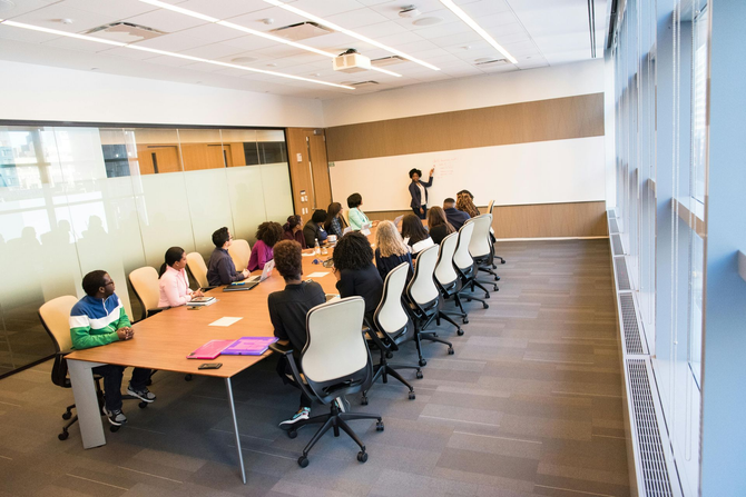 People in a business meeting around a long table; a presenter stands near a whiteboard.