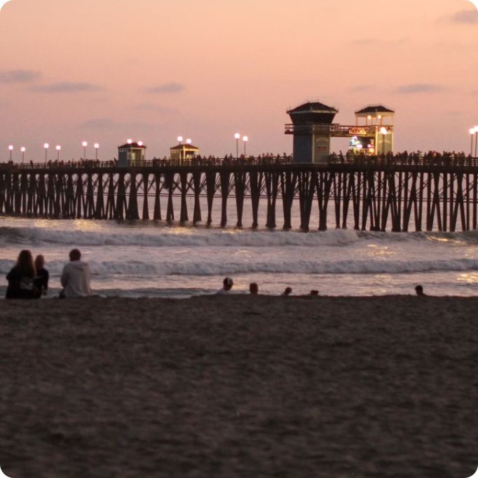 A group of people sit on a beach near a pier