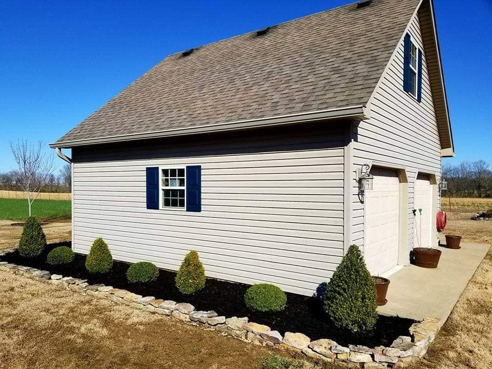 Two-story garage with light siding, dark roof, blue shutters, and manicured landscaping in a sunny field.