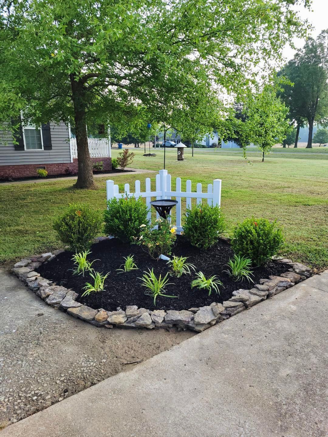Small garden bed with a white picket fence, various plants, black mulch, and stone edging.