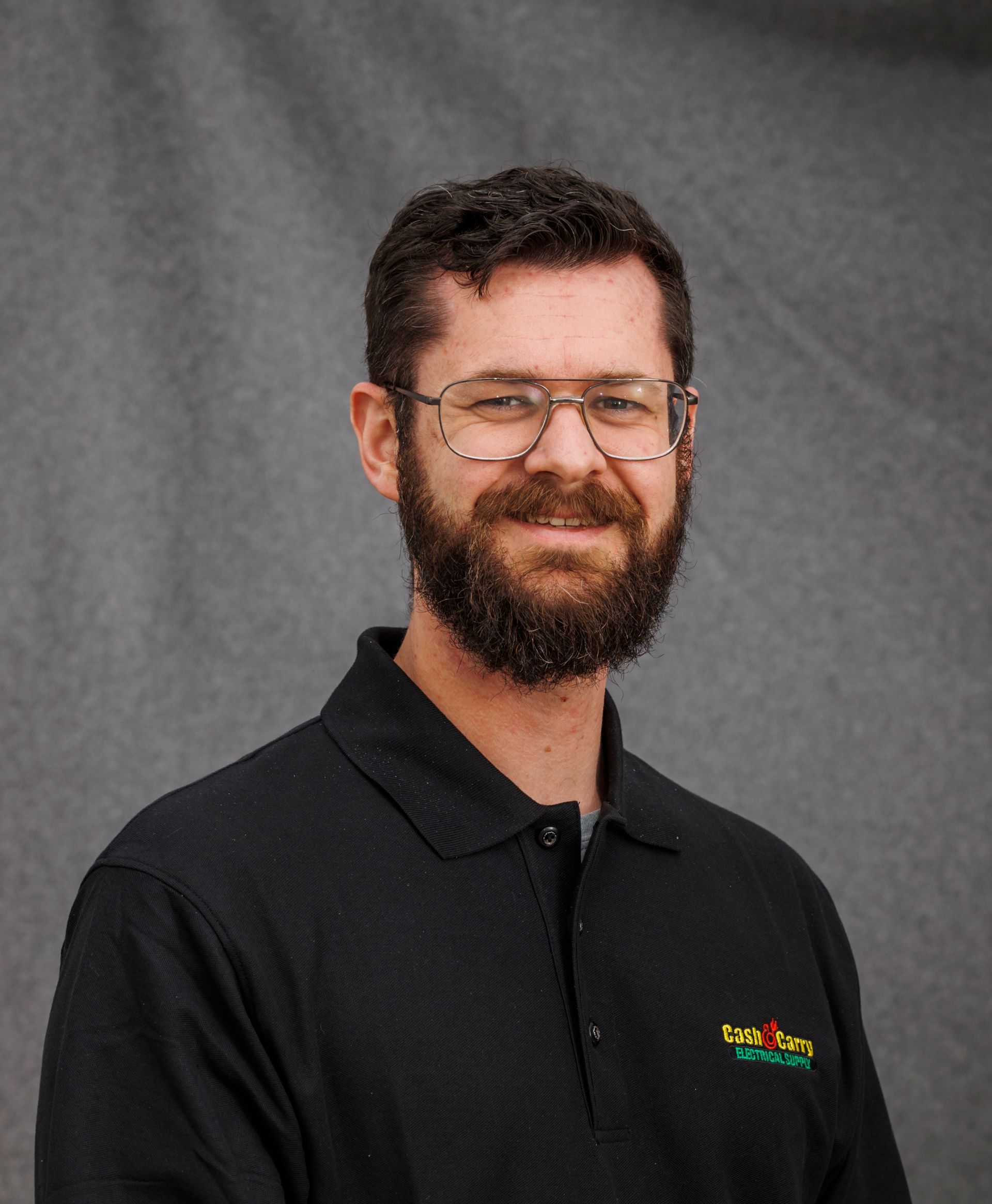 A man with a beard and glasses is wearing a black polo shirt.