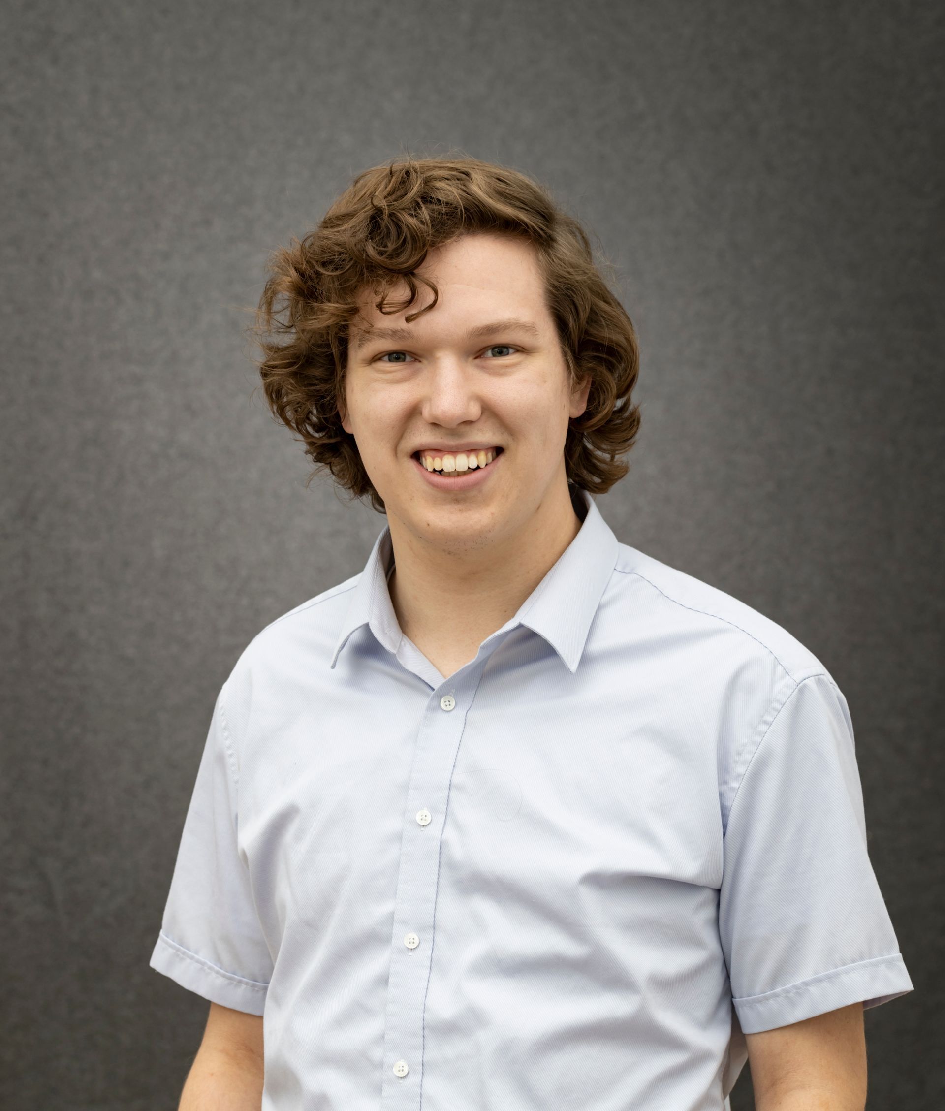 A young man with curly hair is wearing a blue shirt and smiling.