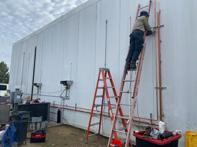 A man on a ladder is working on a pipe on the side of a building.