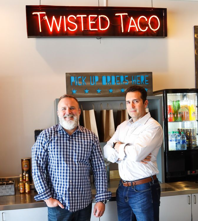 Two men standing in front of a sign that says twisted taco