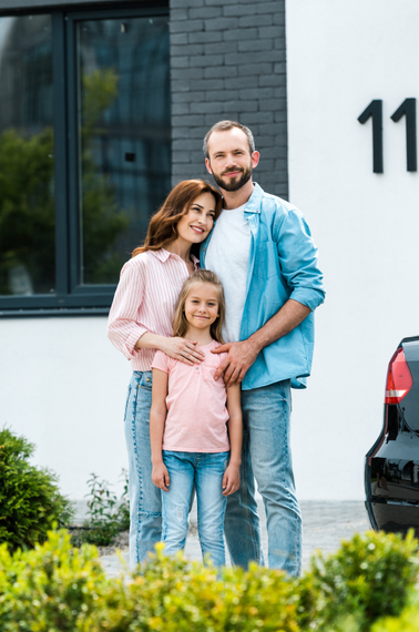 Family of three standing outside a white building, smiling and embracing near a black car.