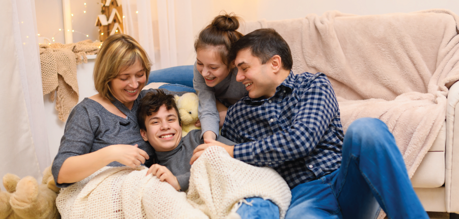 Four friends cuddle on a couch, smiling and relaxing under a blanket in a cozy living room.