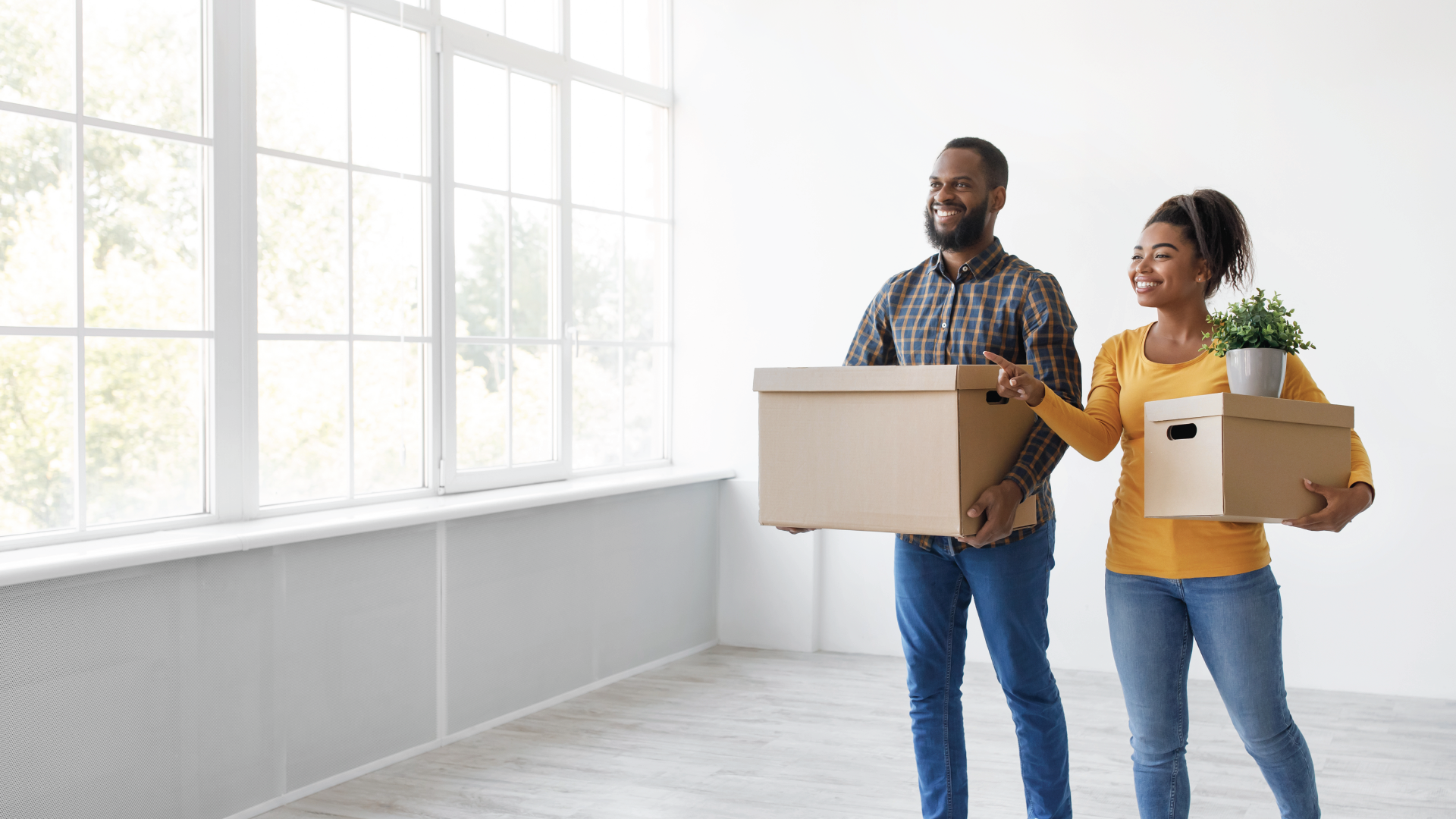 Two people carrying moving boxes in a bright, empty room with large windows
