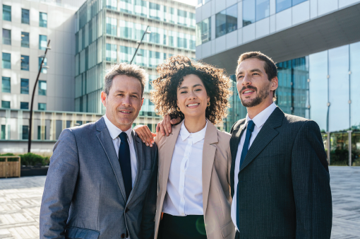 Three business professionals standing outside modern office buildings, smiling at the camera.