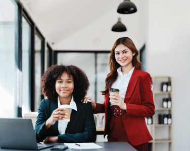 Two coworkers in a bright office, one seated with a coffee and one standing beside her smiling.