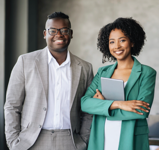 Two smiling professionals in business attire stand side-by-side, one holding a tablet, against a neutral office background.