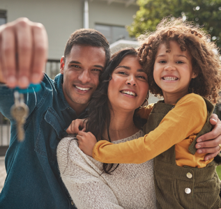 A smiling family holds up a house key in front of their home, celebrating a new residence.