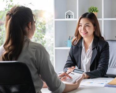 Two women in a bright office discuss finances, one holding a calculator and papers across a desk.