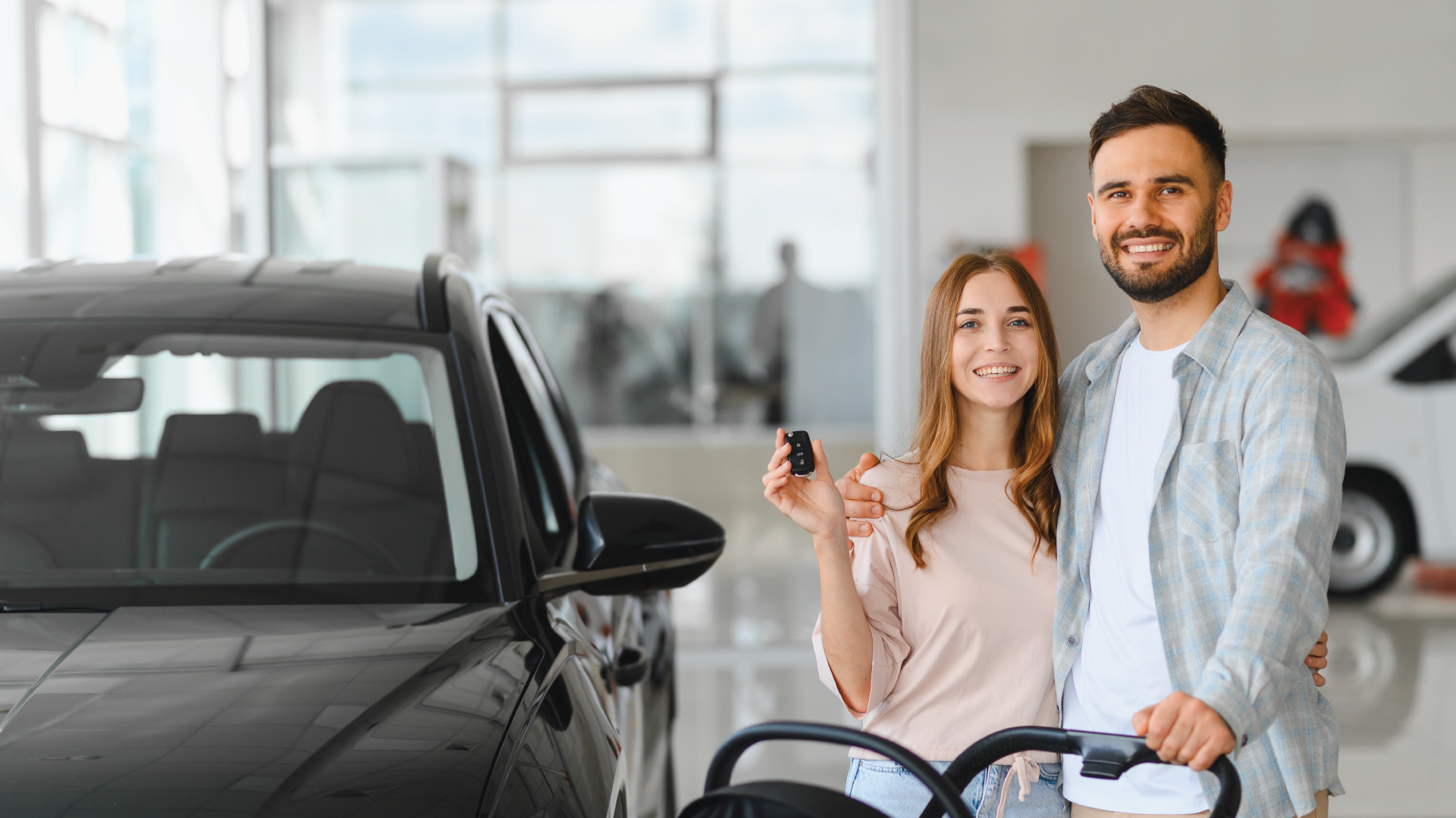 A couple smiles while holding car keys next to a new black car in a dealership showroom.