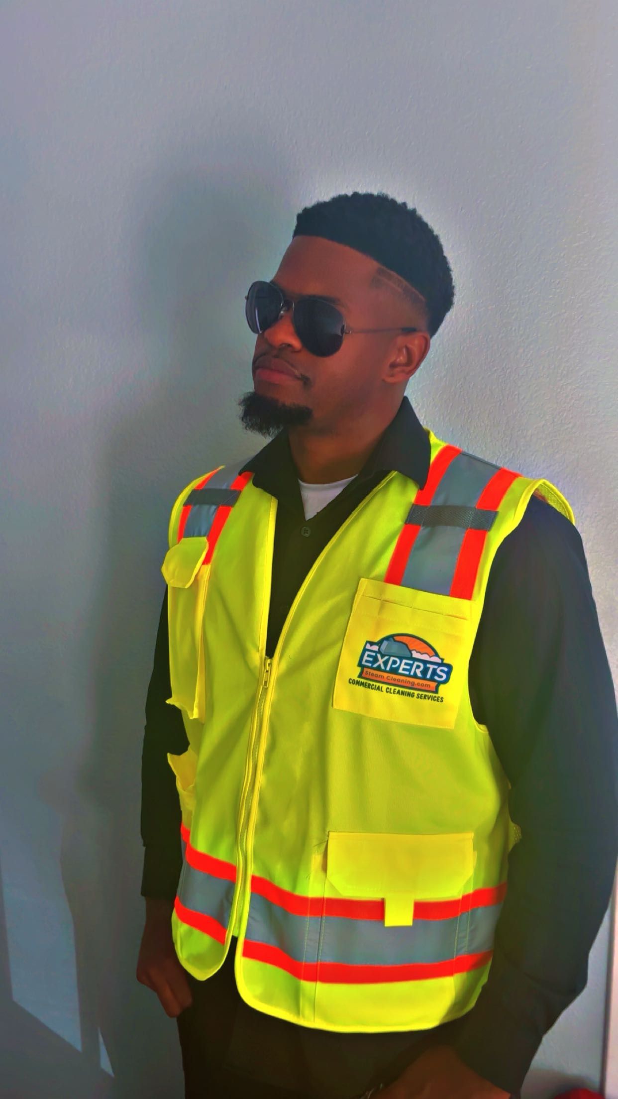 Person wearing a reflective safety vest and sunglasses, standing against a neutral-colored wall.