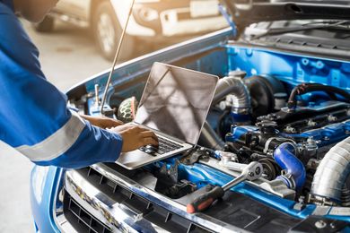 Mechanic using a laptop to diagnose a blue car’s open engine bay in a garage