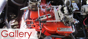 Red racing engine with chrome carburetors and red hoses in a garage display, labeled “Gallery”