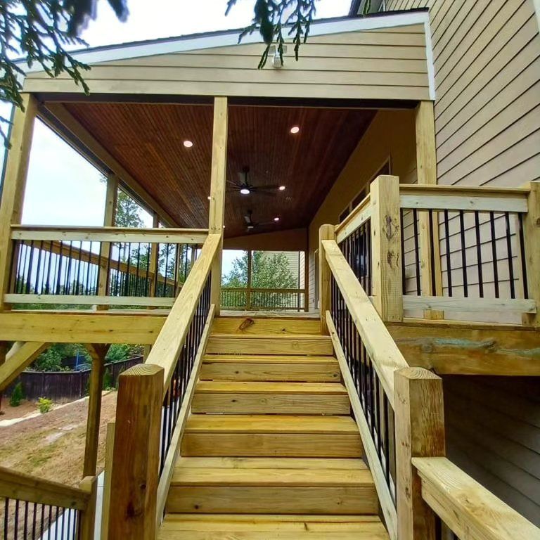 Wooden deck with stairs leading up to a covered porch with dark railing and ceiling fan.
