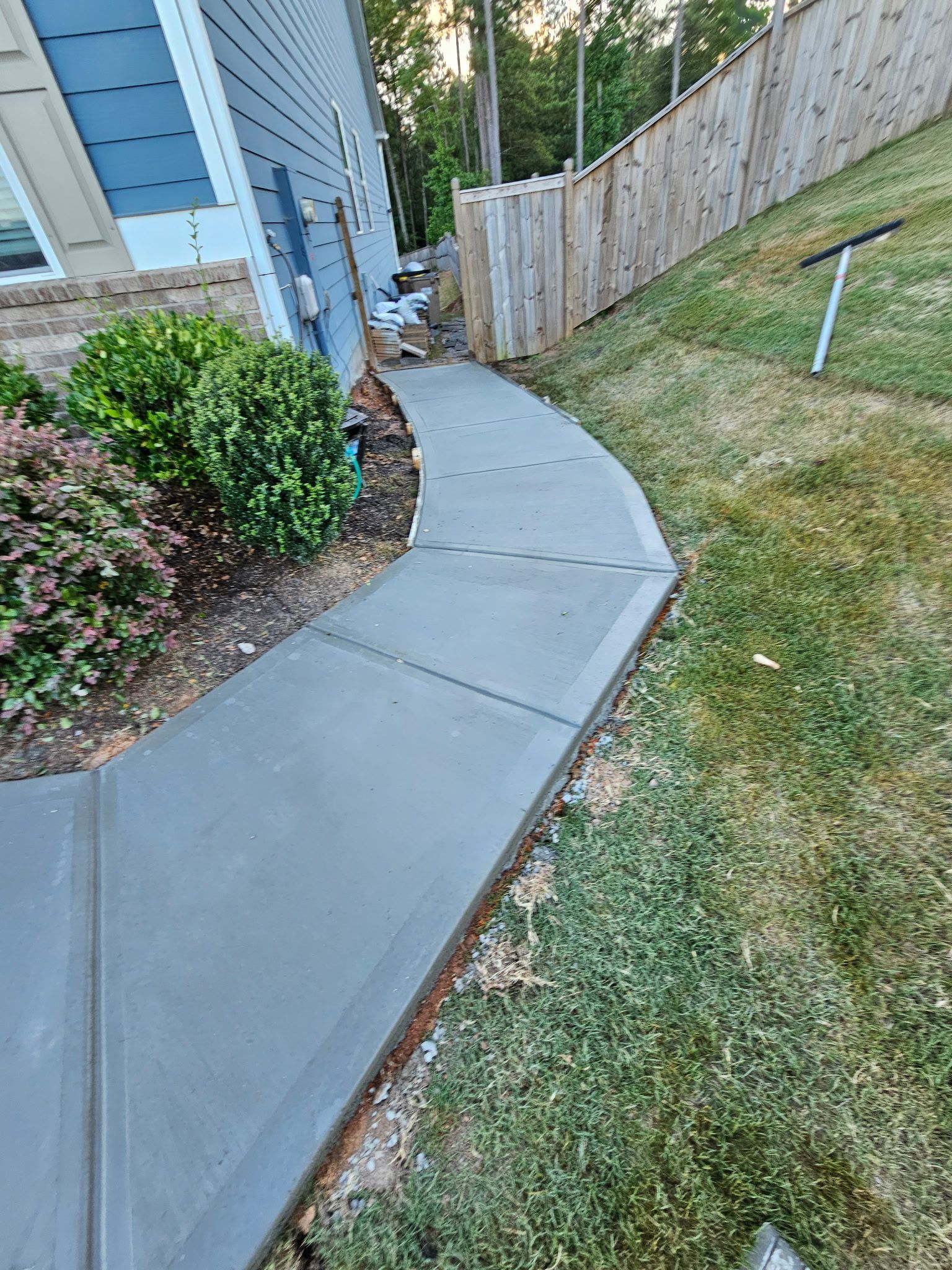 Newly poured concrete walkway next to house and lawn, leading toward a wooden fence.
