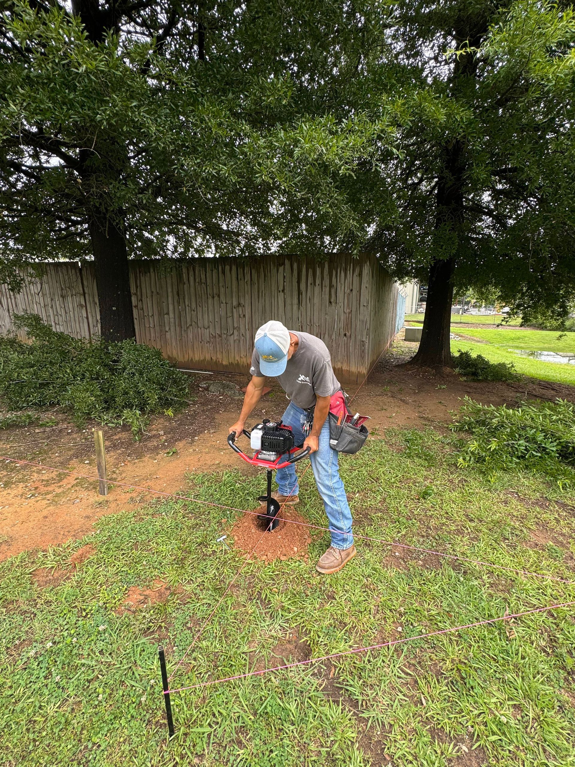 Man using a drill to make a hole in the ground in a grassy area near trees.