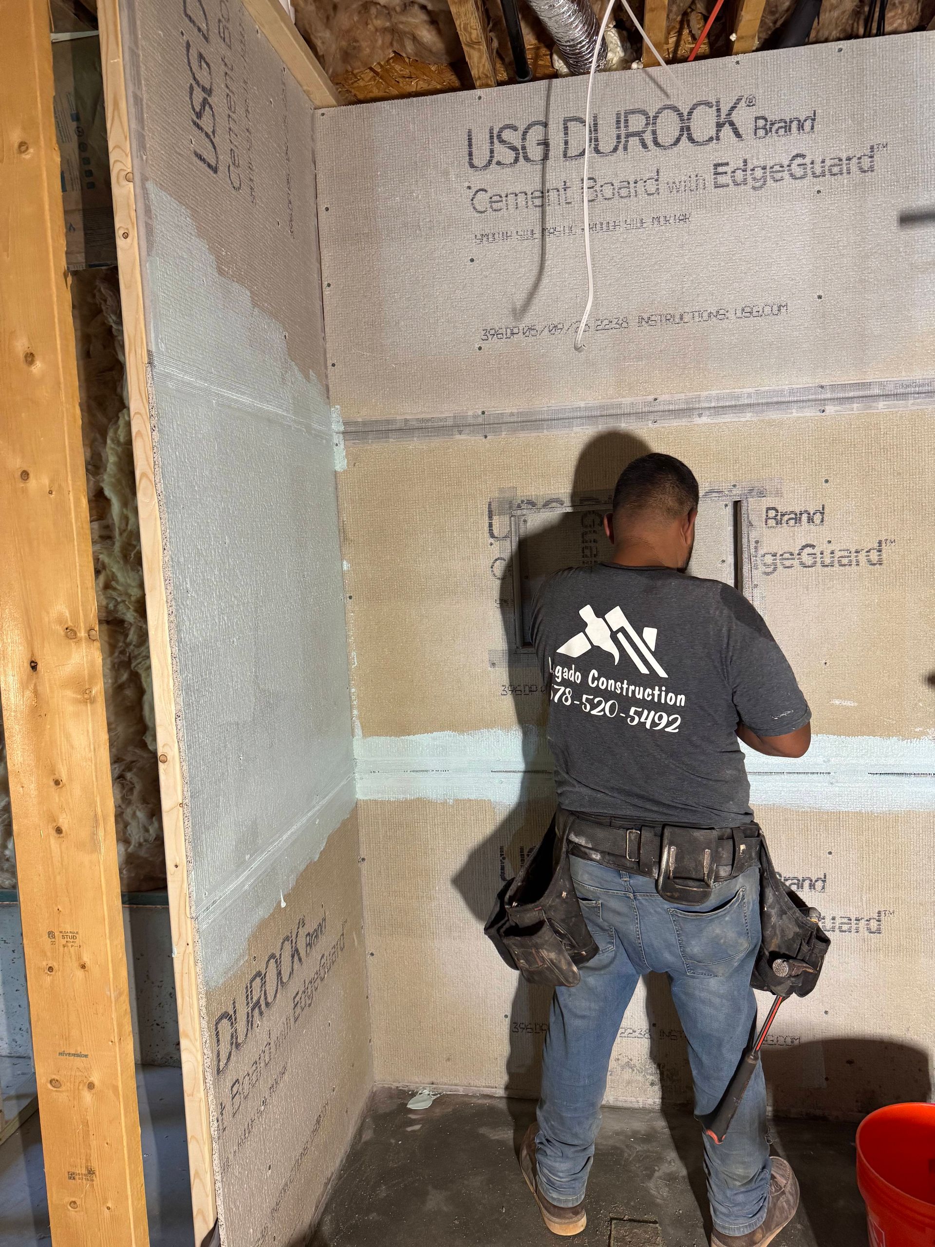 Man installing cement board in a shower stall. The walls are gray, with visible tape and text.