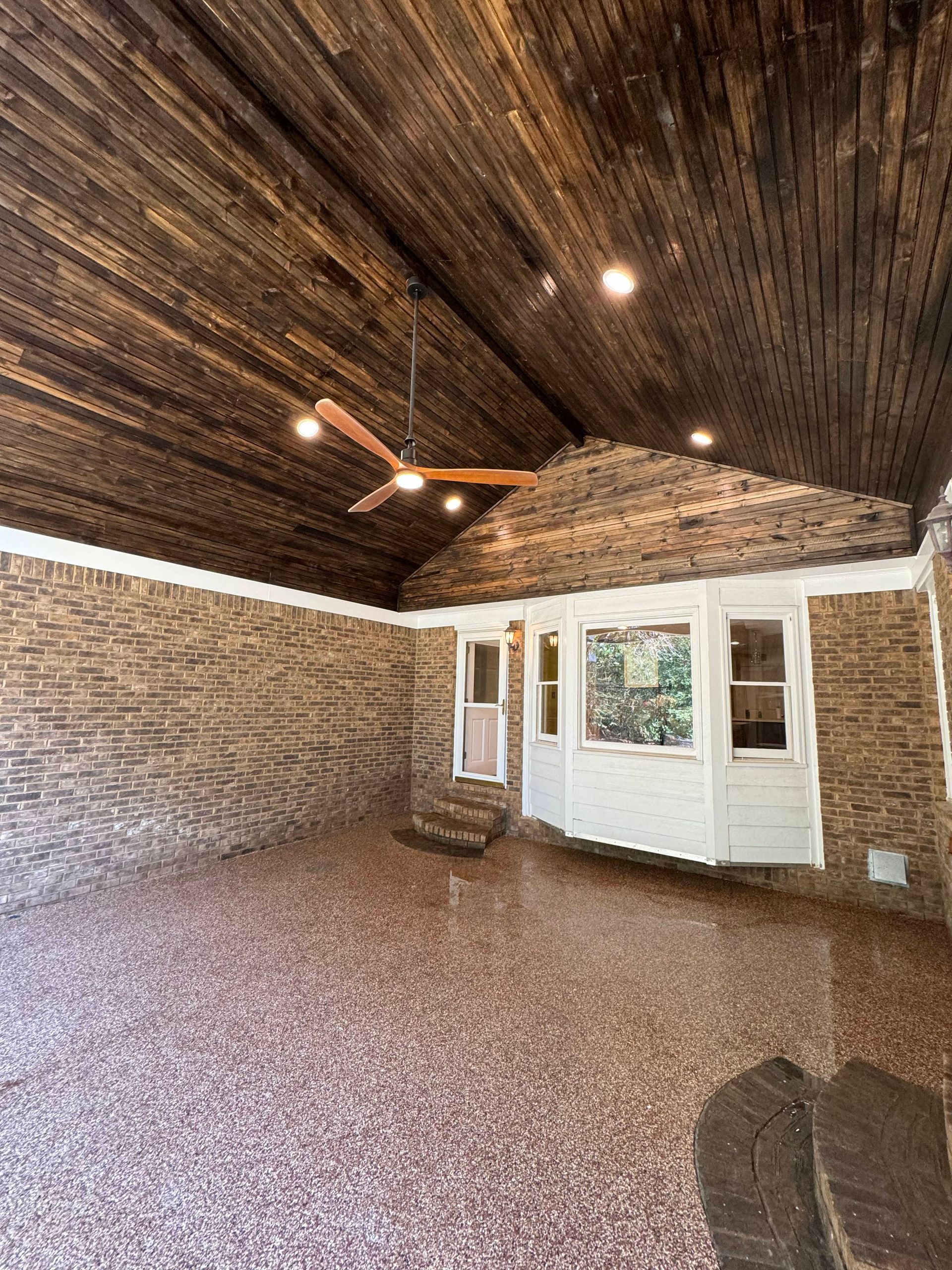Covered outdoor patio with wood ceiling, brick walls, and pebble floor. A ceiling fan and windows are visible.