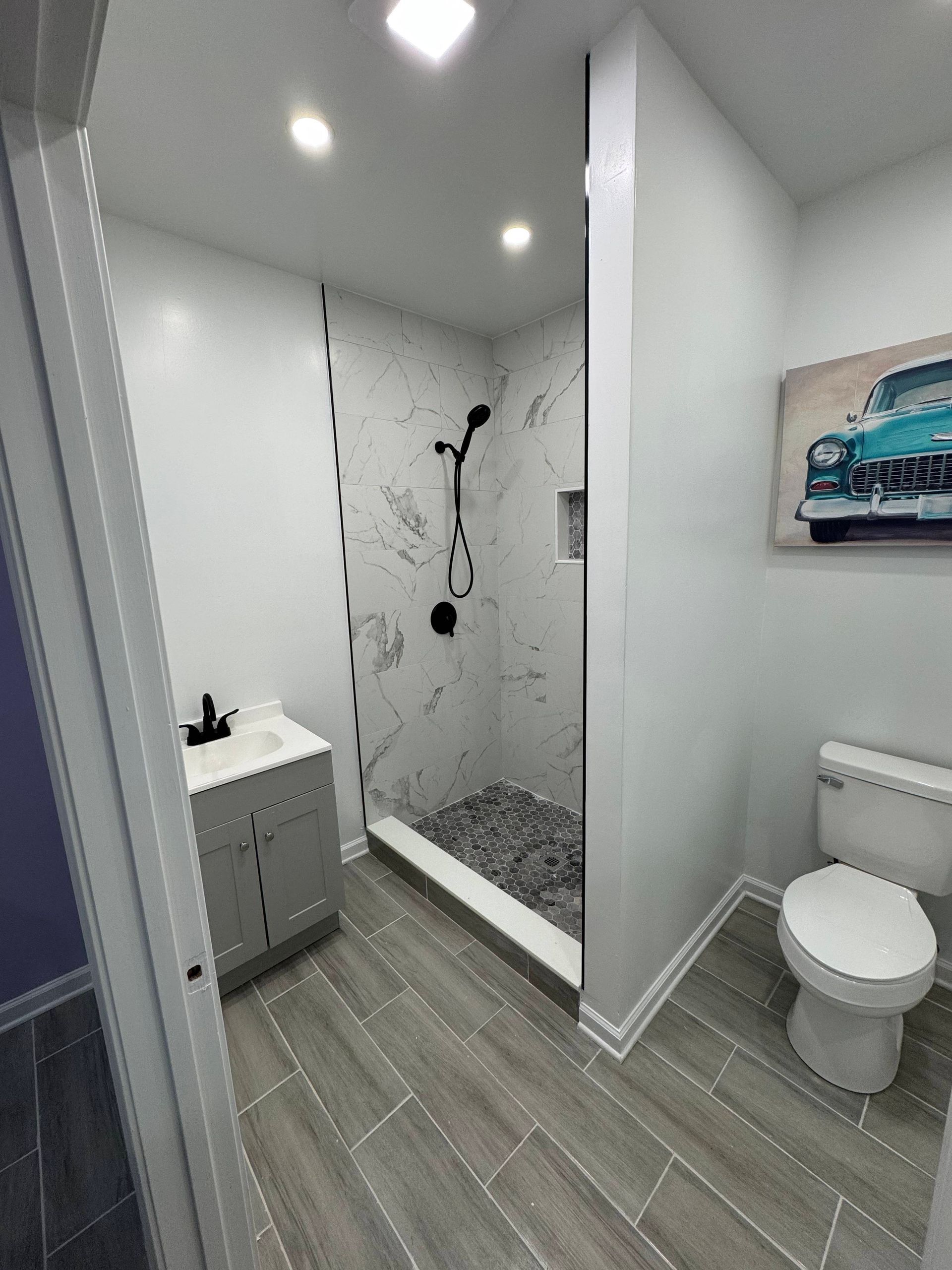 Modern bathroom with gray and white tile, black showerhead, toilet, and vanity. A vintage car painting hangs on the wall.
