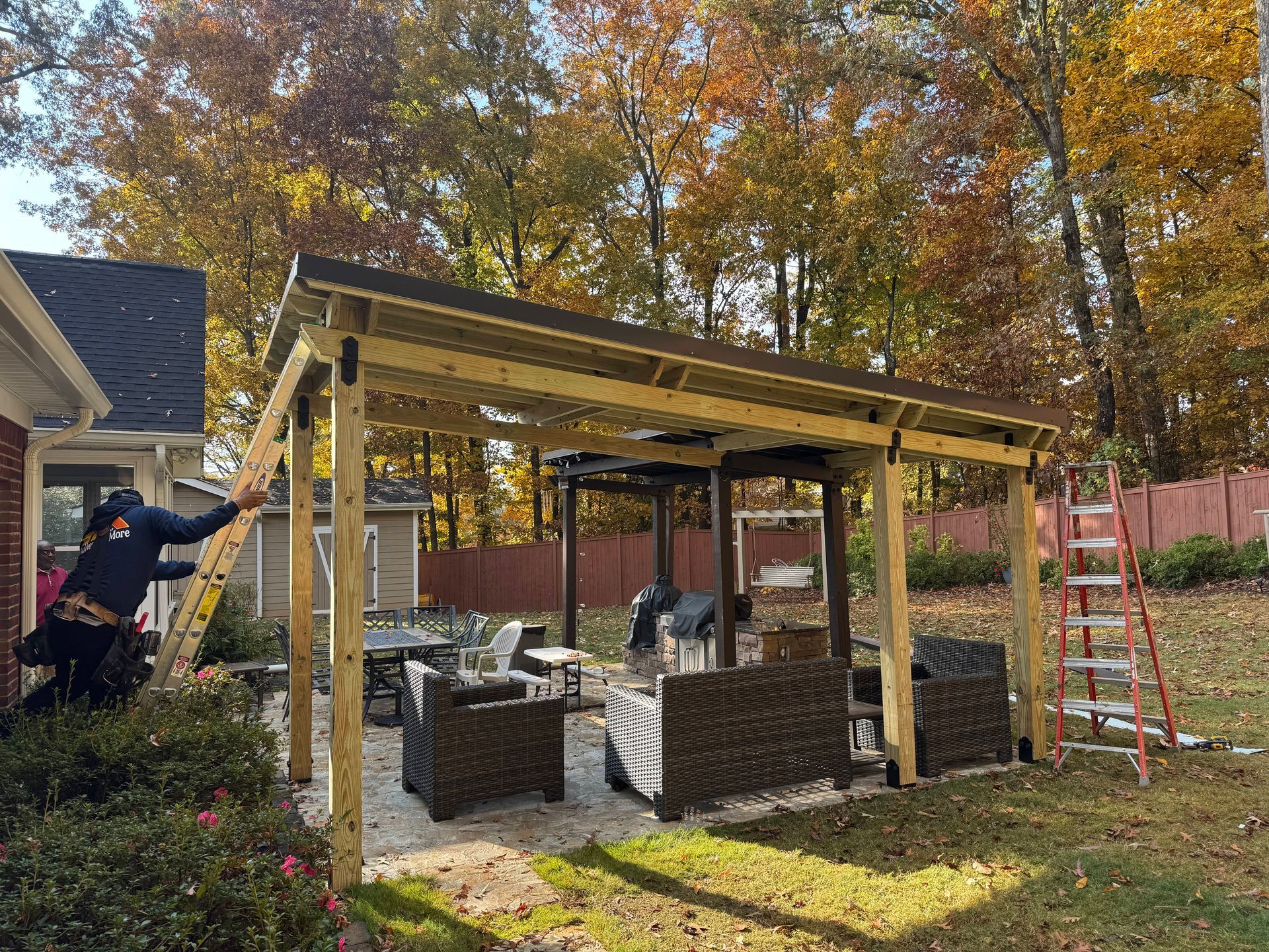 Man on ladder installing a wooden patio cover over outdoor furniture. Autumn leaves in the background.