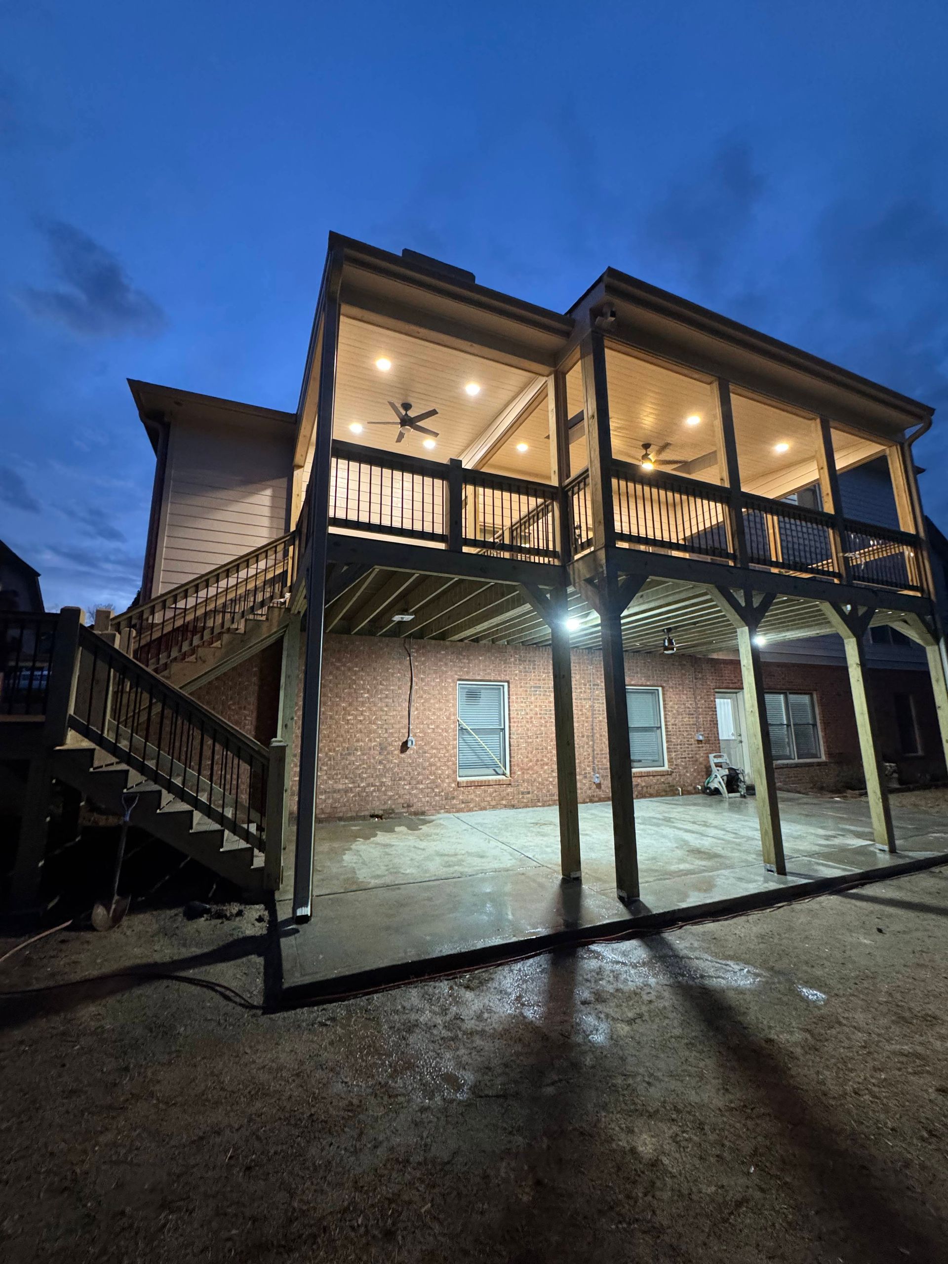Two-story house with a wooden deck and brick foundation, illuminated at dusk.