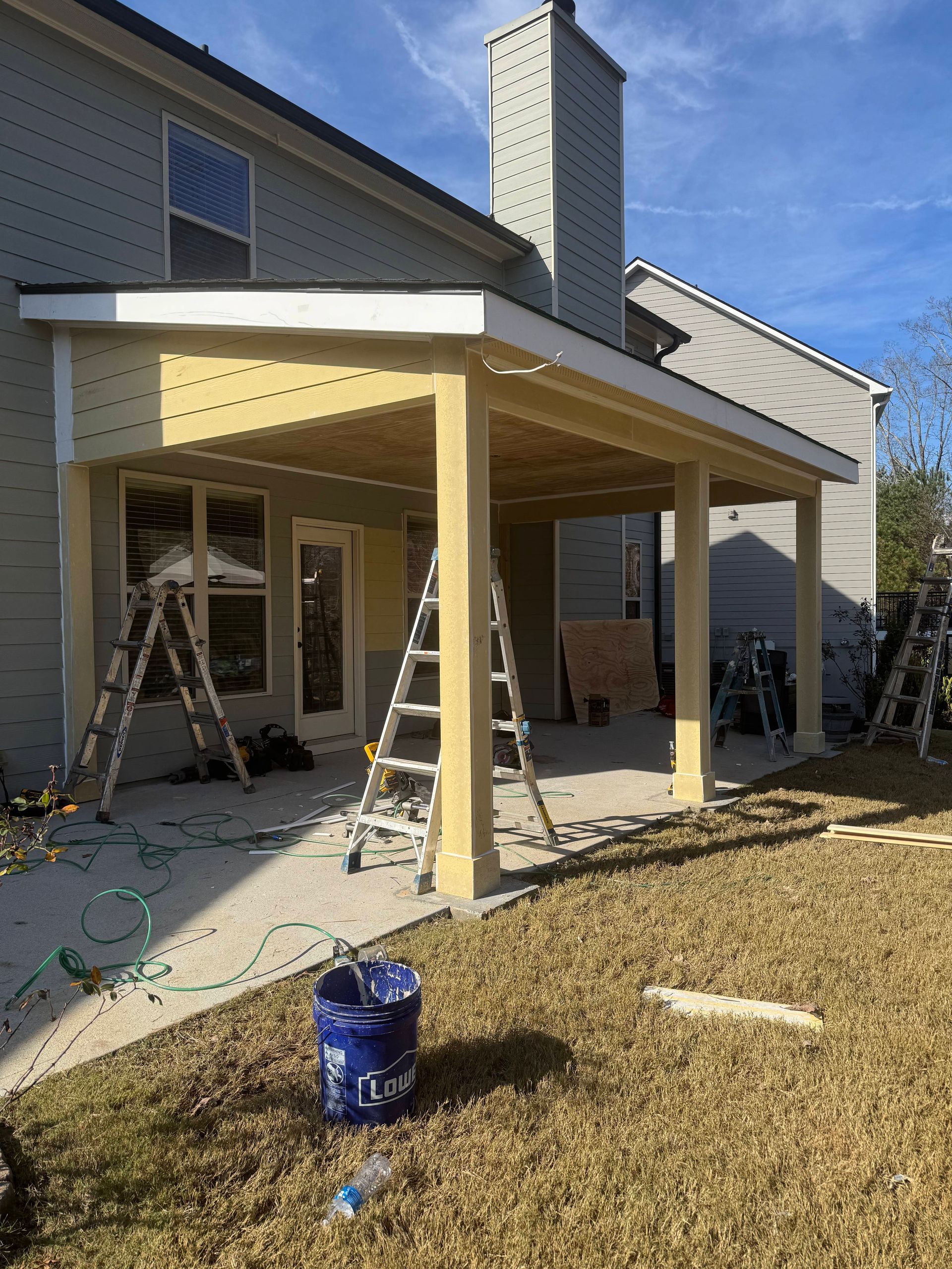 Construction of a covered patio with tan columns and roof, ladders, and tools on a sunny day.