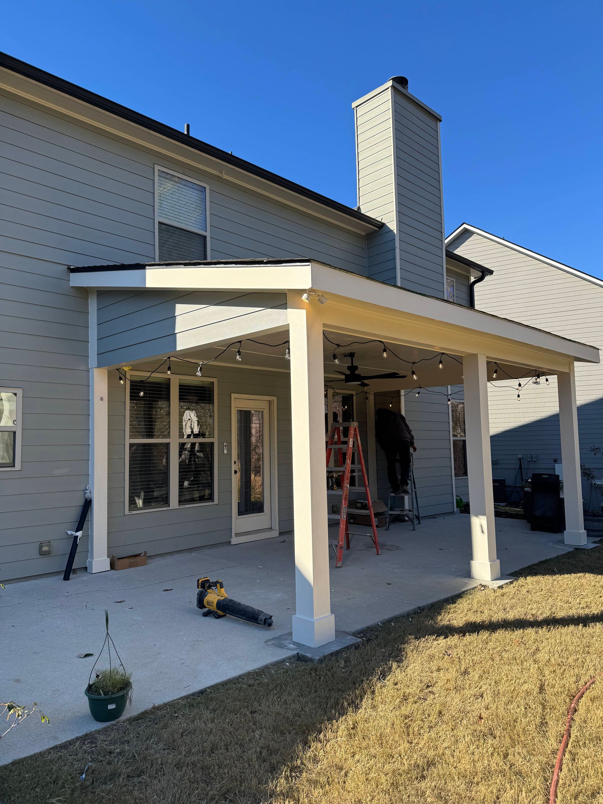 A light-colored patio cover attached to a gray house, with a concrete patio and yard.