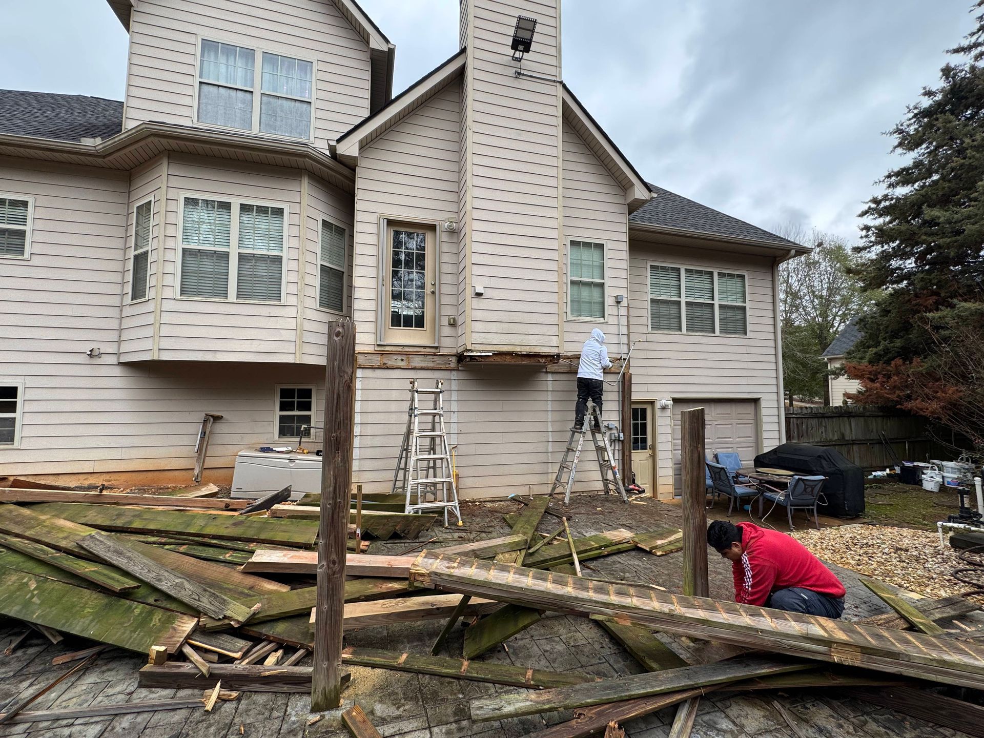 Deck demolition in progress at a beige house. Workers remove boards; debris litters the ground.