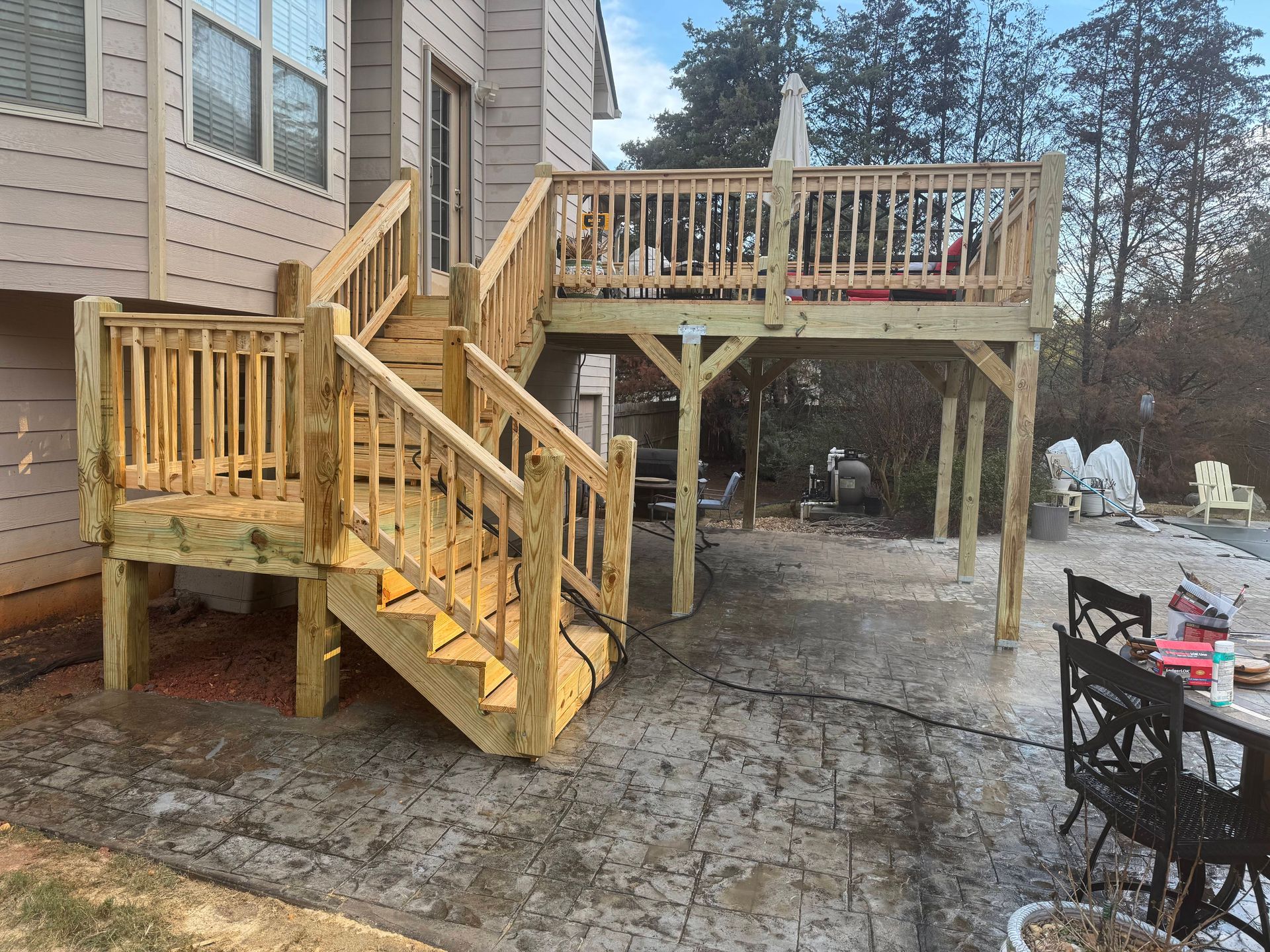 Wooden deck with stairs, extending from a house to a concrete patio. Trees and sky in the background.