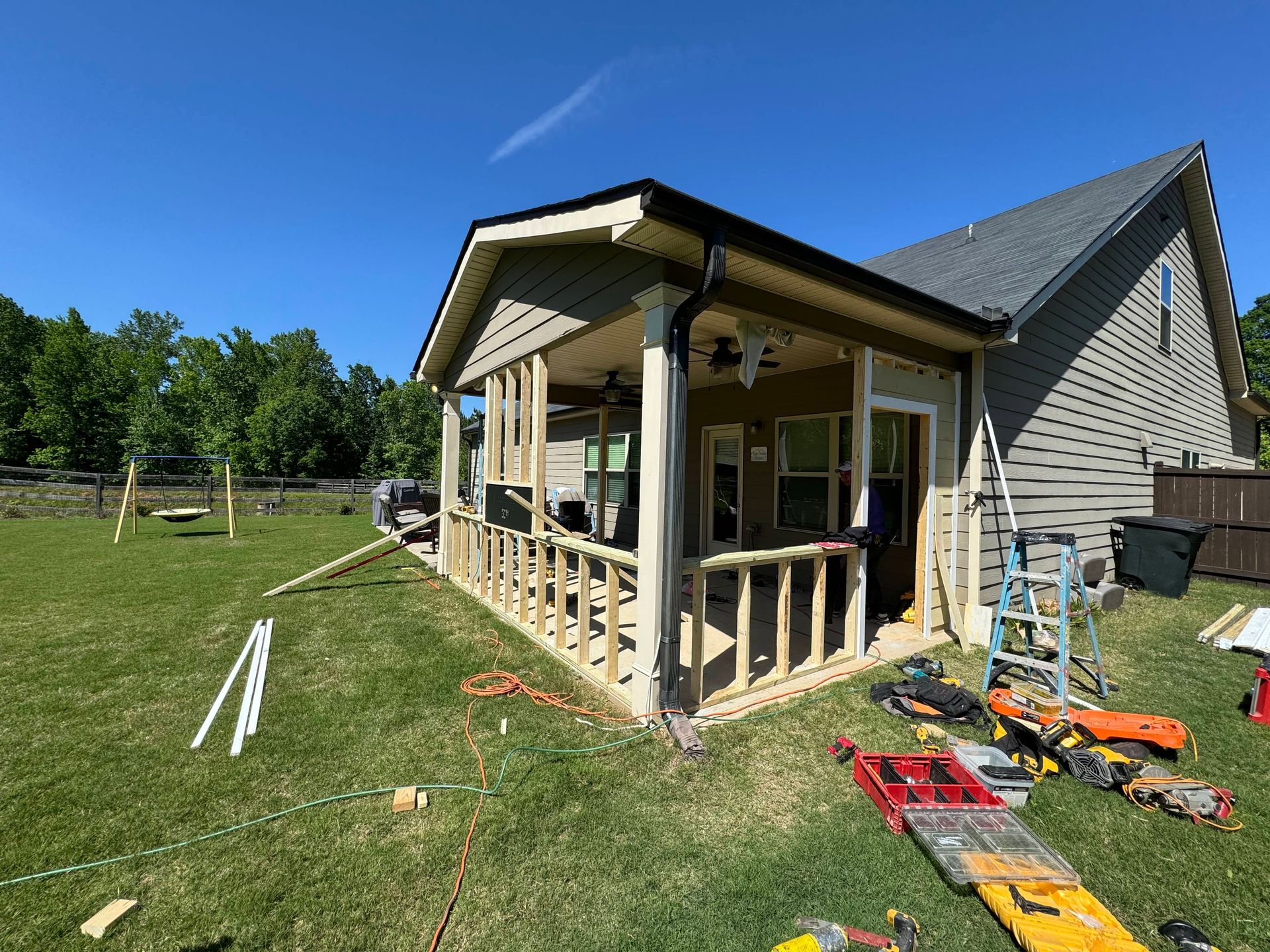 A new porch under construction with exposed framing, beige siding, and tools scattered on the grass.