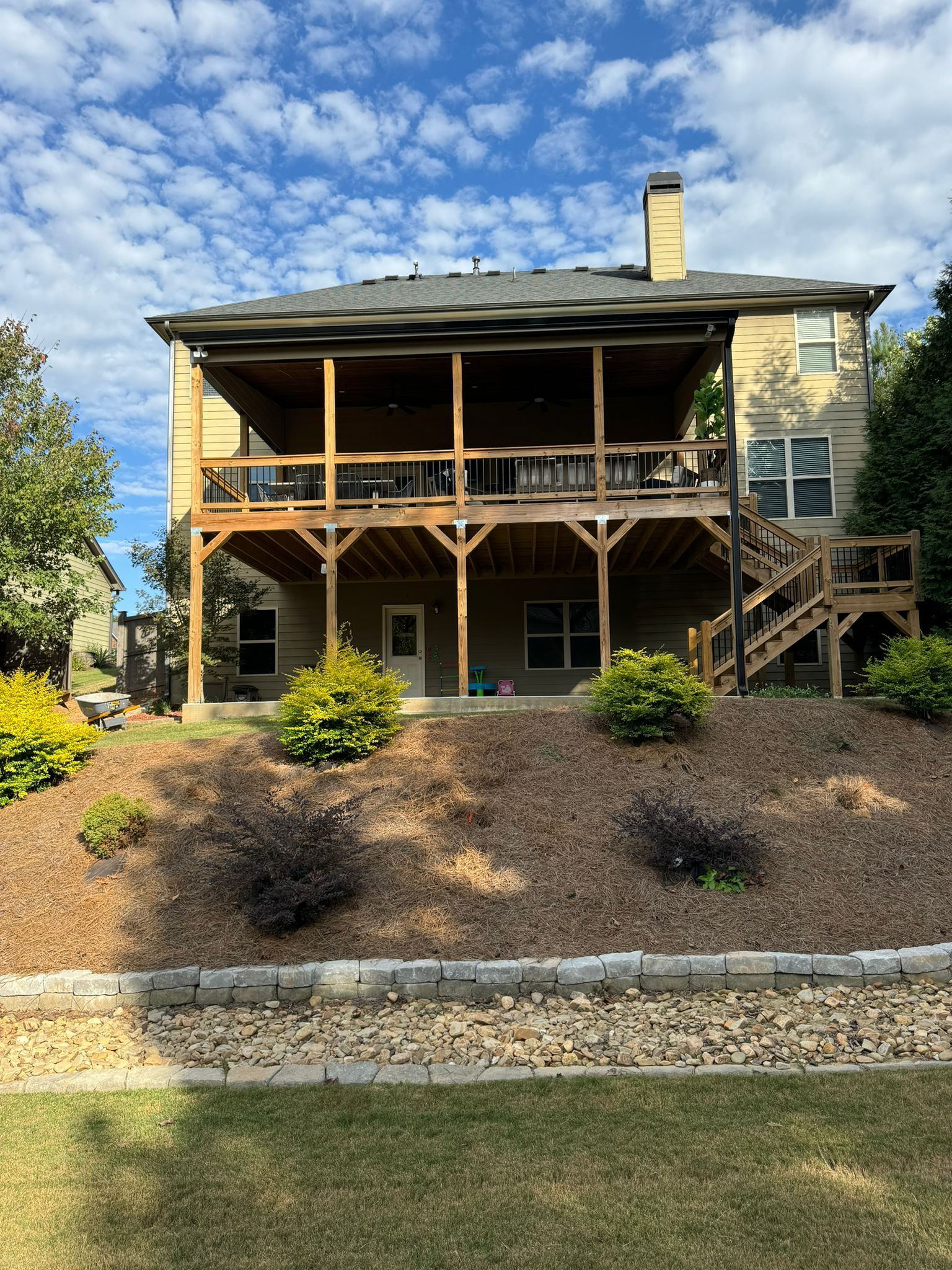 Two-story house with a wooden deck and covered porch. Landscaping includes mulch, bushes, and a retaining wall.