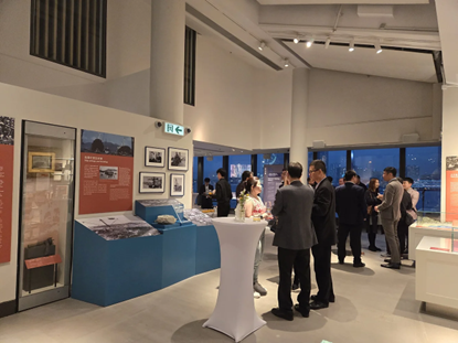 People gather at an exhibition in a brightly lit room with display cases. Some are conversing near a small table.