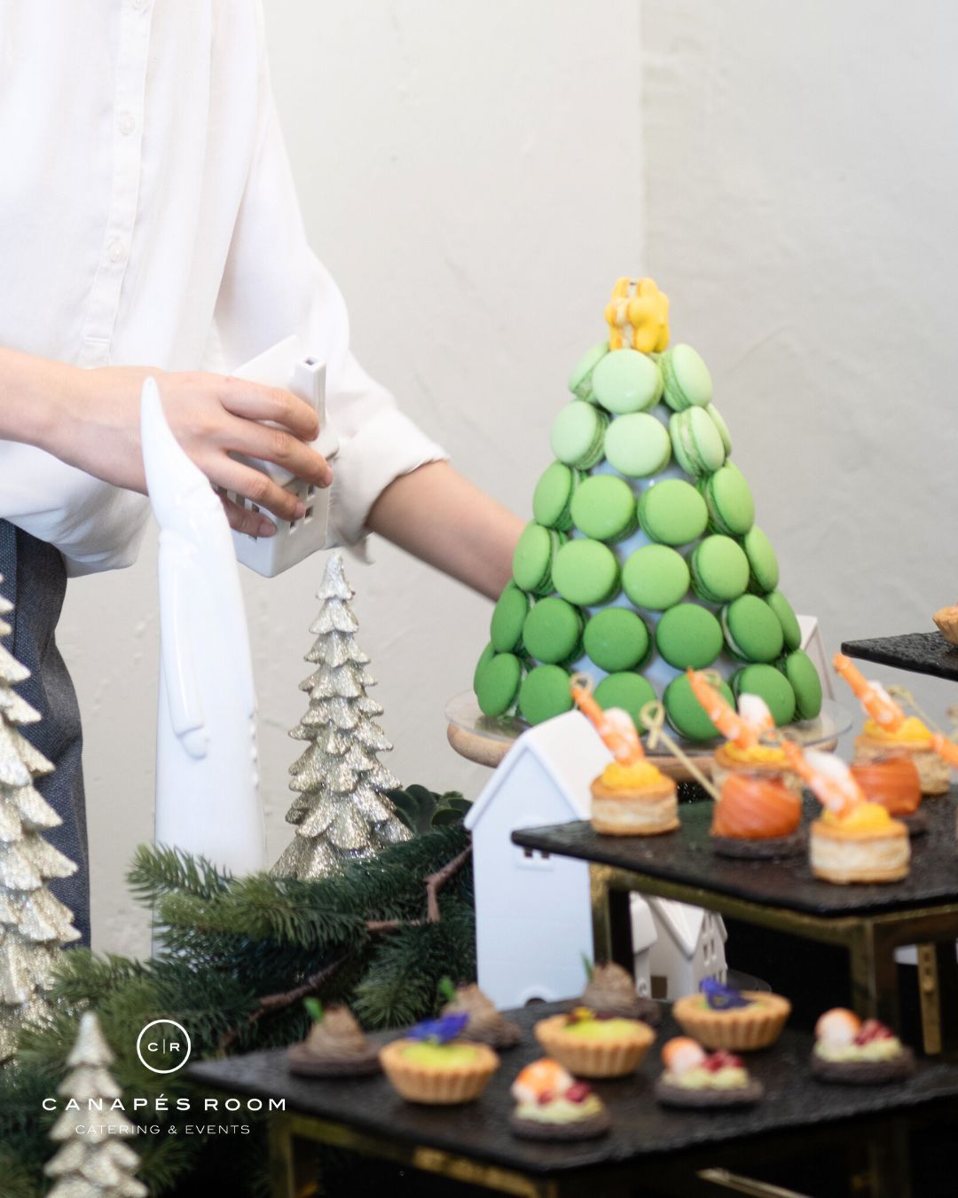 Person arranging macarons shaped as a Christmas tree, with other appetizers on tiered serving platters.