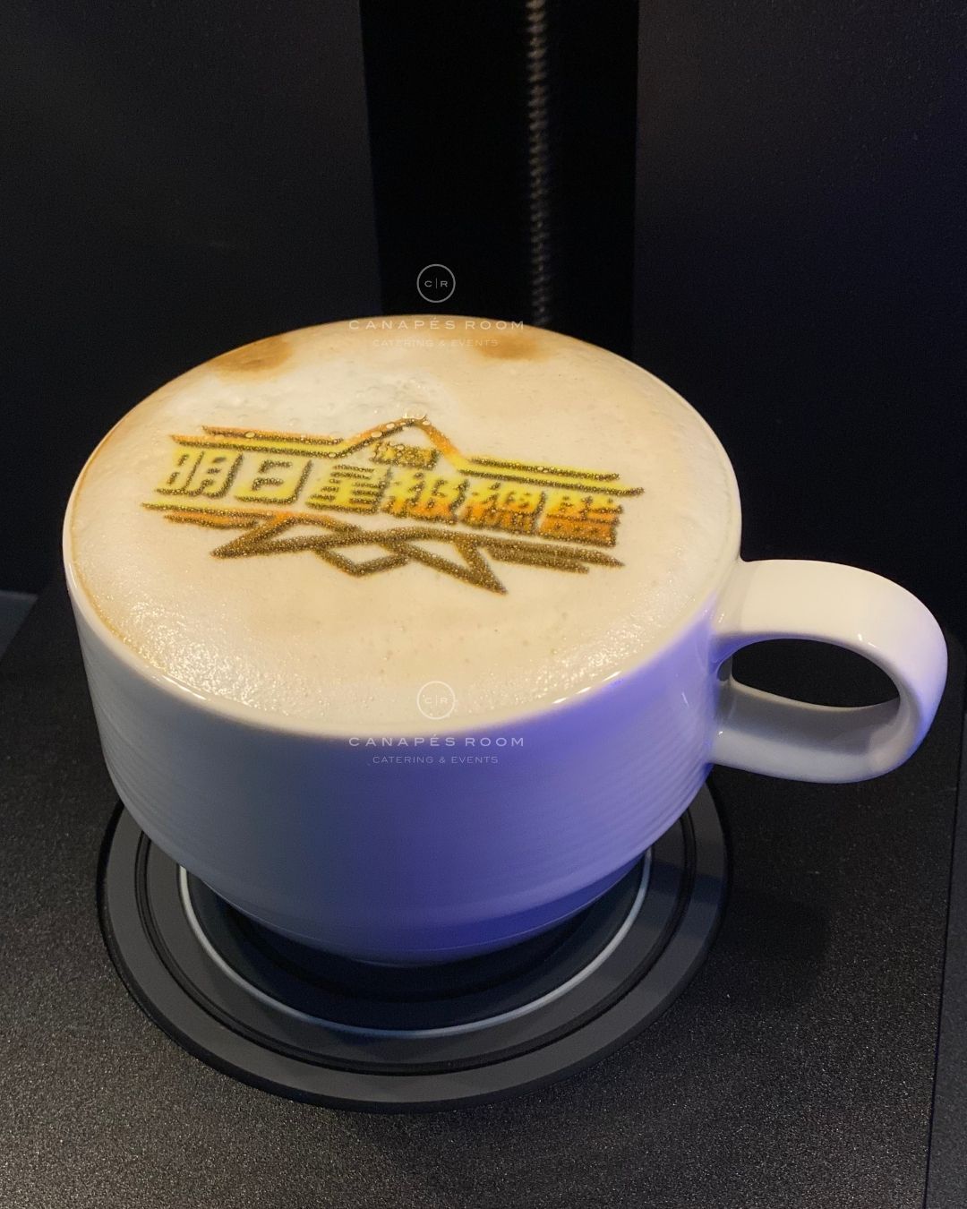 拿鐵盛在一個白色馬克杯裡，泡沫上印著馬克杯的標誌。馬克杯放在一個黑色杯墊上。