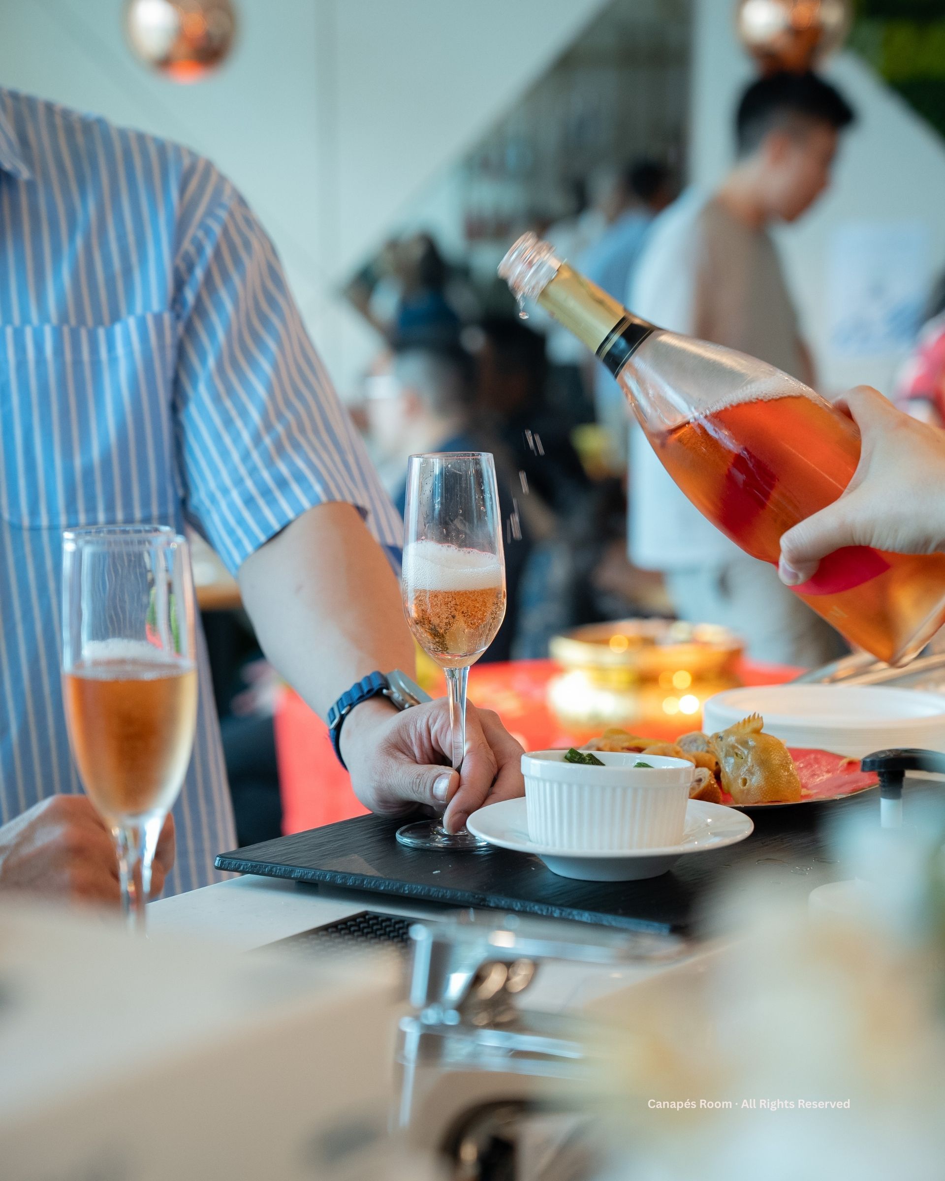 一個人將香檳倒入玻璃杯中。背景是桌上擺放的開胃菜和其他客人。