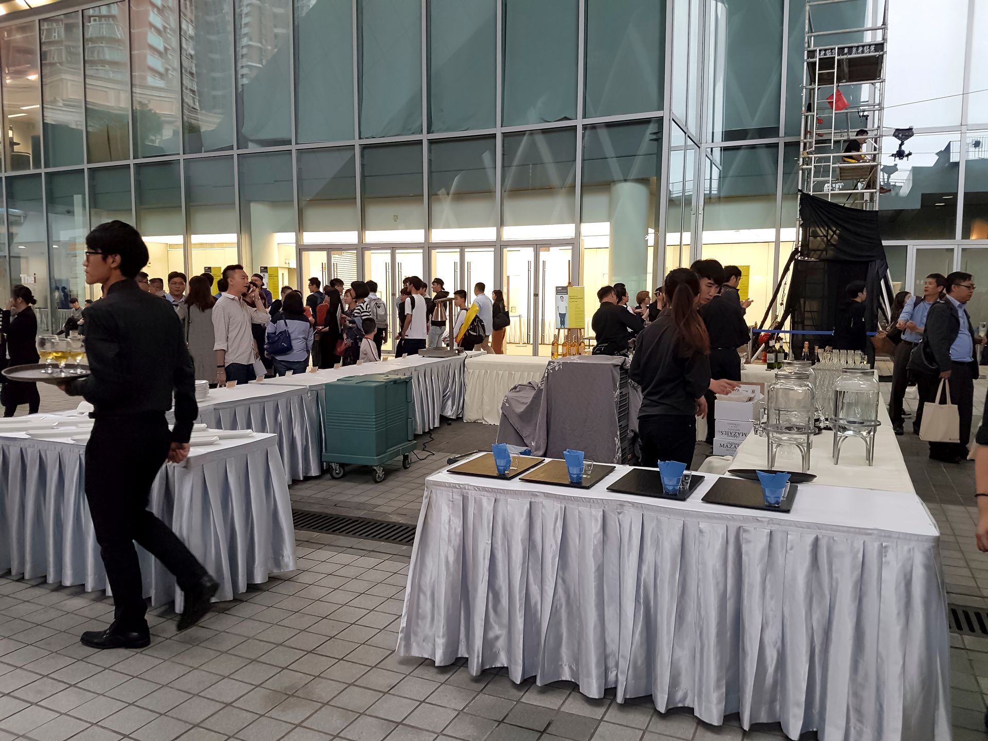 Event with food and drink tables; servers in black uniforms. People gathered near a glass-walled building.