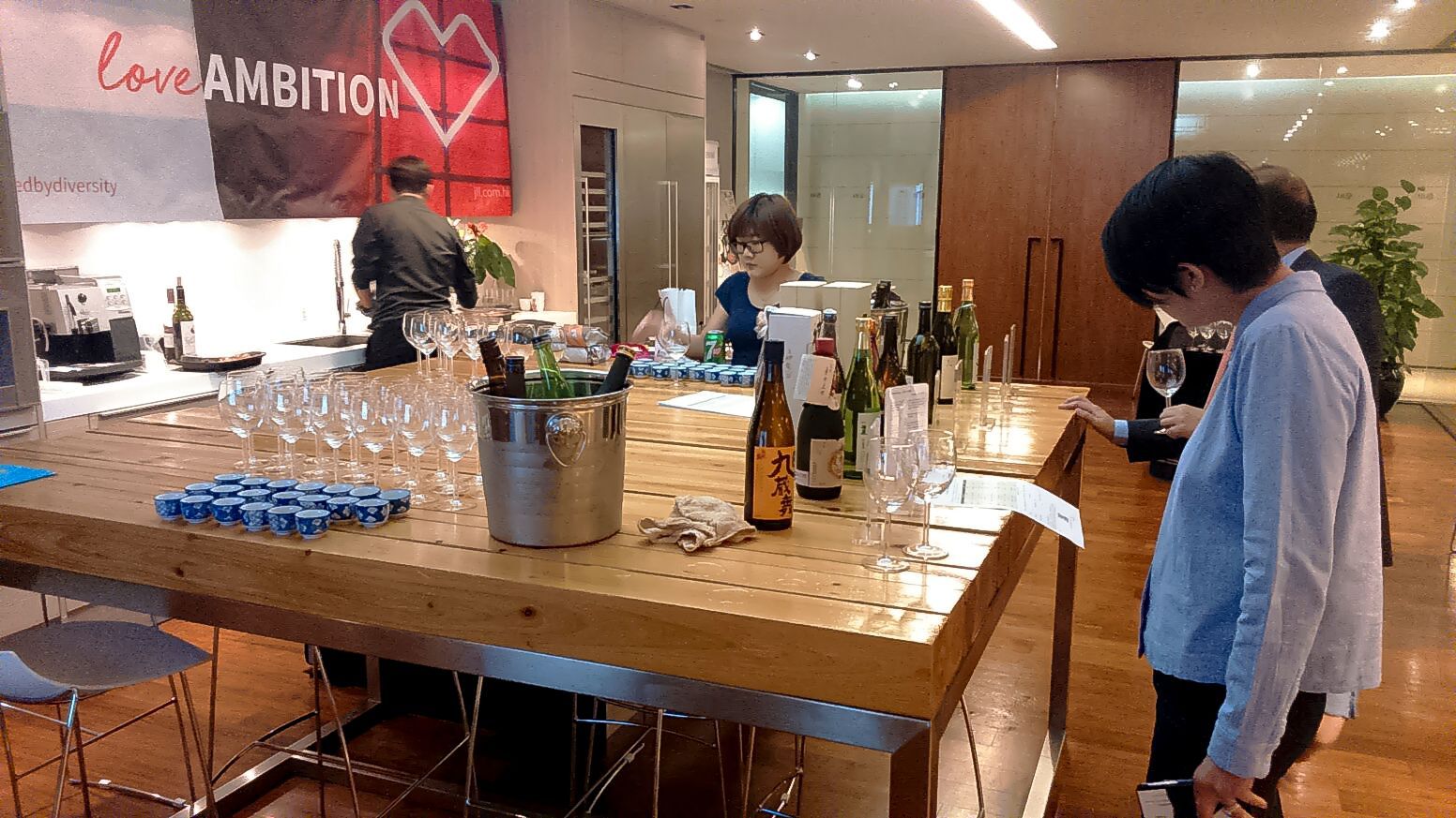 People at a bar, some looking at bottles of wine, ice bucket.