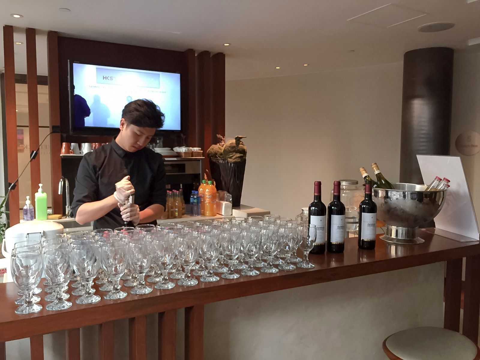 Bartender mixing a drink behind a bar, many glasses lined up, bottles of wine, TV in the background.