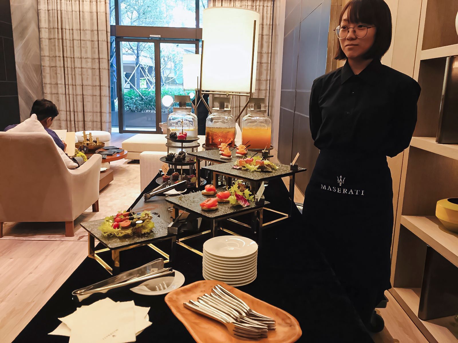 Woman in black uniform stands near a buffet table with drinks, snacks, and serving utensils.