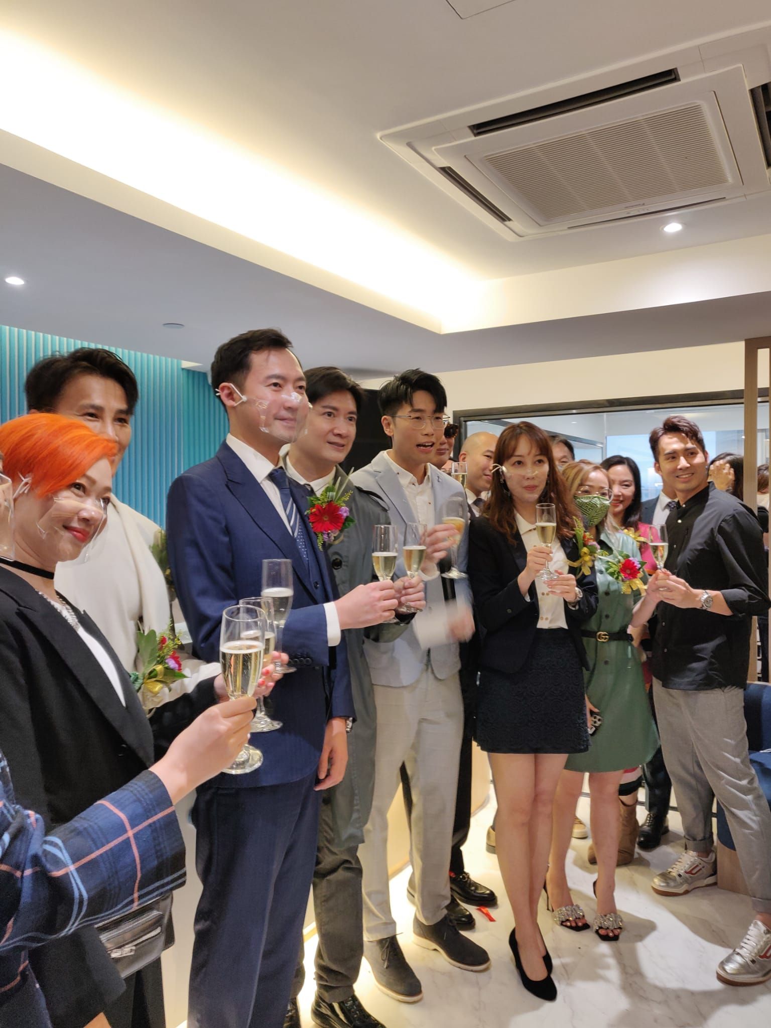 People toasting with champagne in a well-lit interior. Several hold flowers, dressed in suits and business attire.