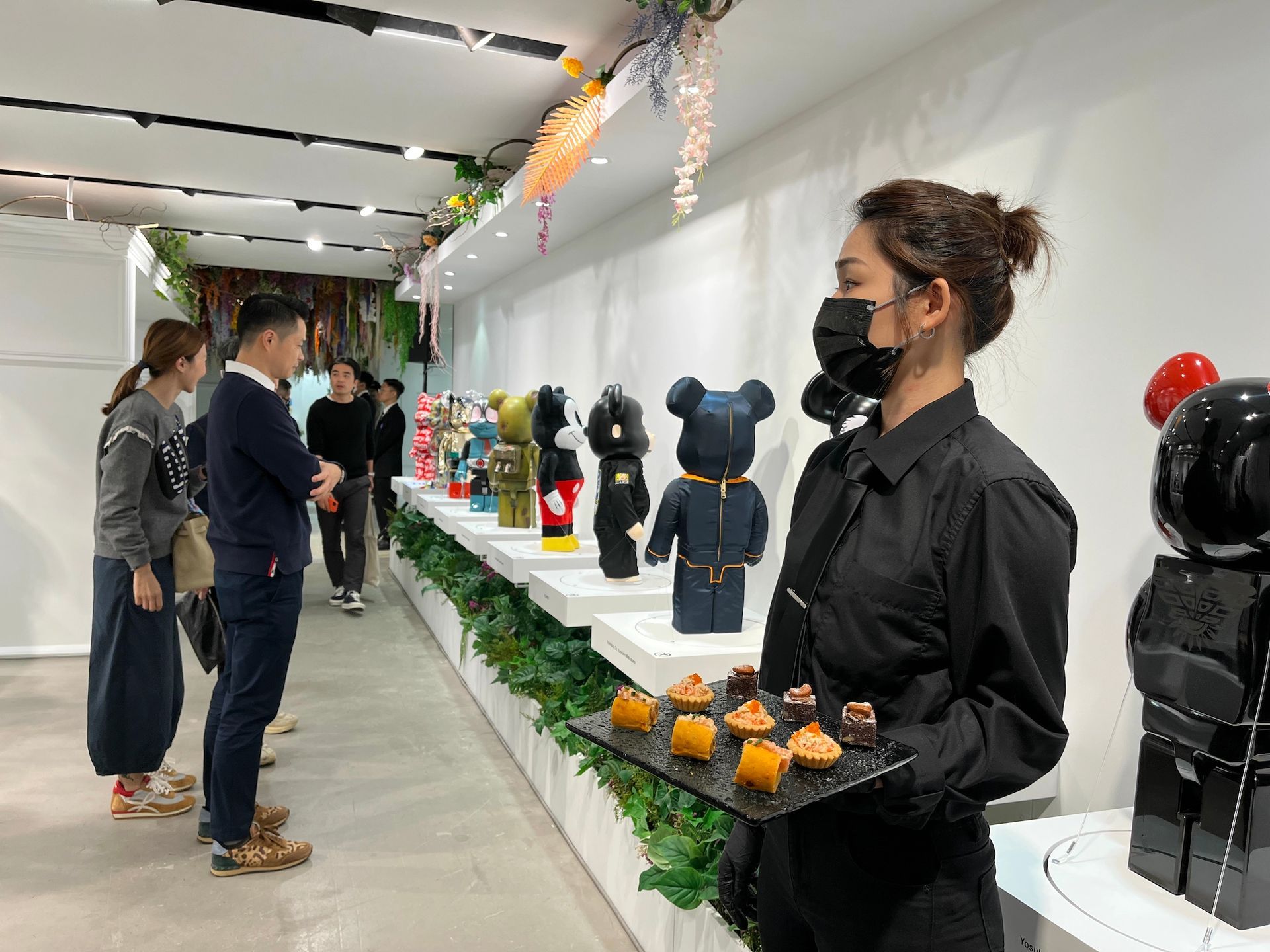 People viewing art in a gallery. A server offers treats. Rows of bear-shaped figures are displayed on white shelves.