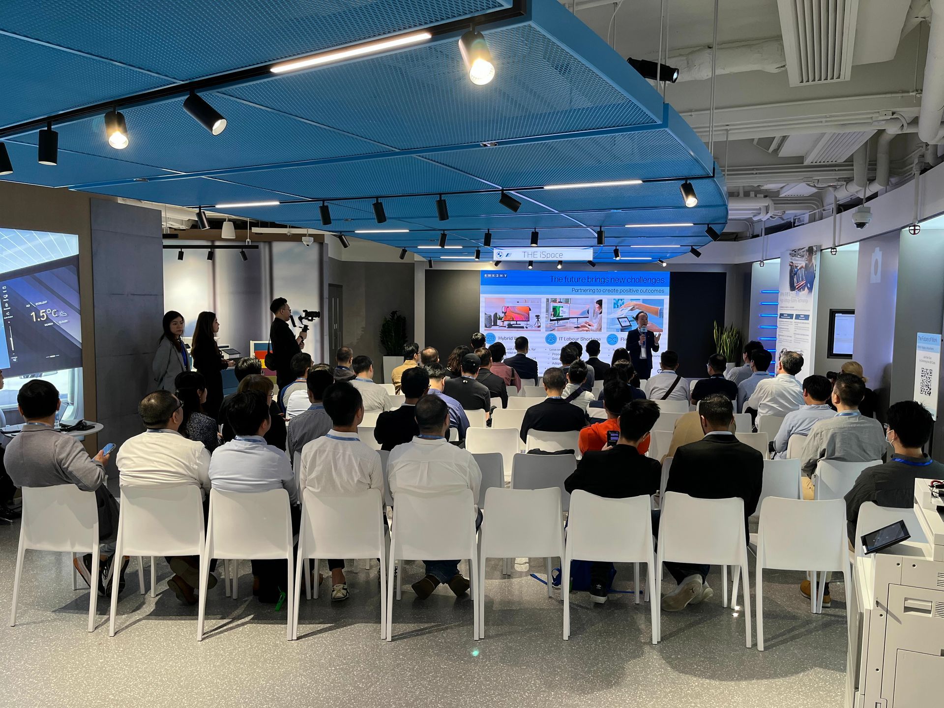 Audience at presentation in modern room with blue accents and large screen.