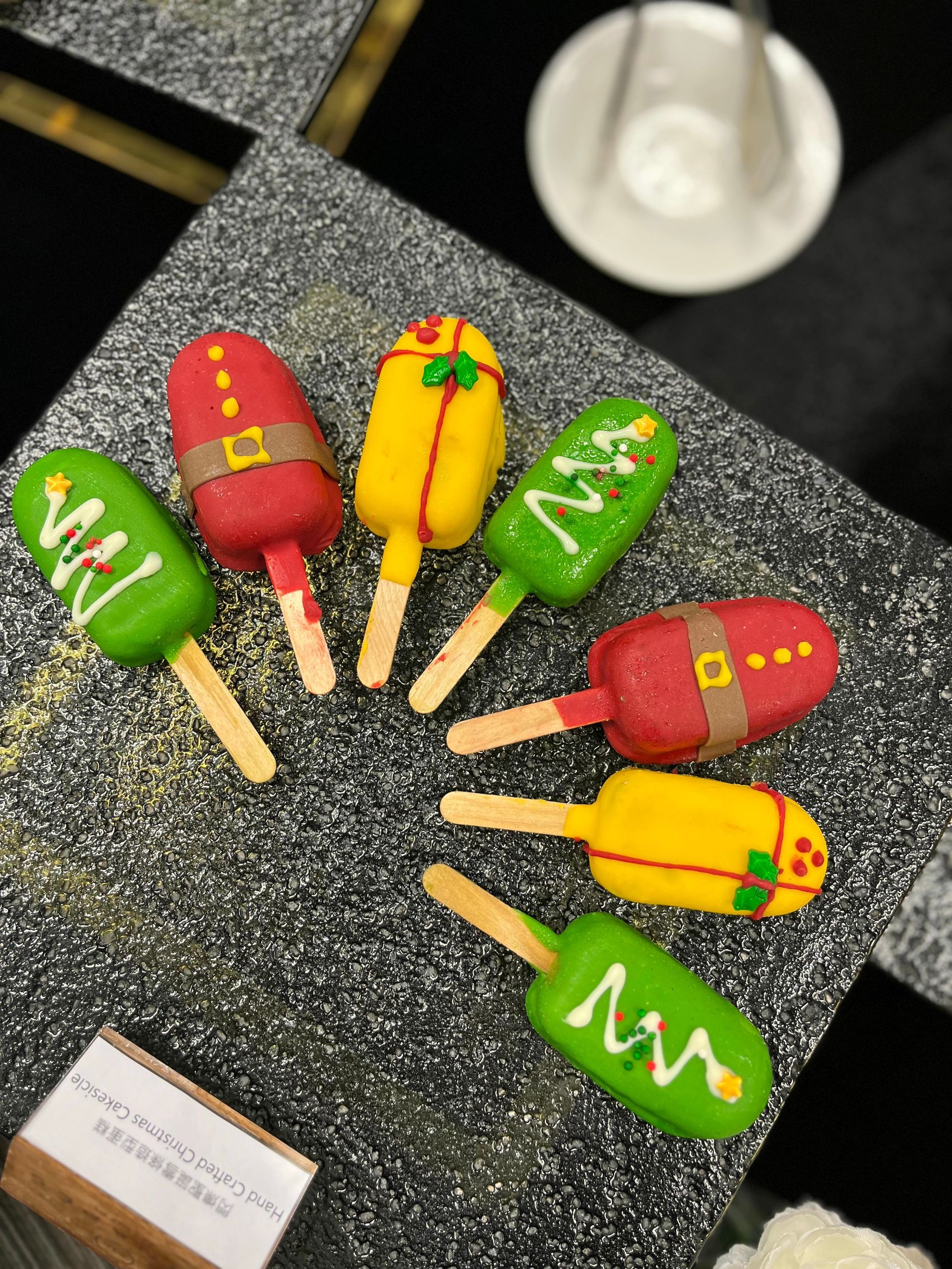 Christmas-themed cake pops on sticks, decorated as trees, Santas, and presents; set on a speckled gray surface.