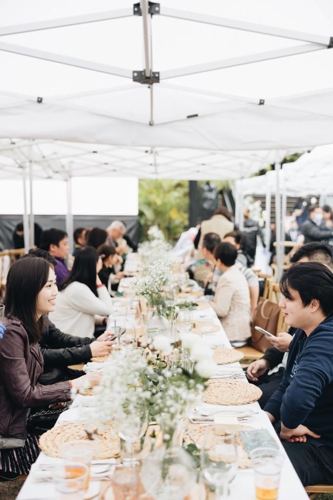 People seated at a long table outdoors under a white tent, eating and talking. Flowers and place settings adorn the table.