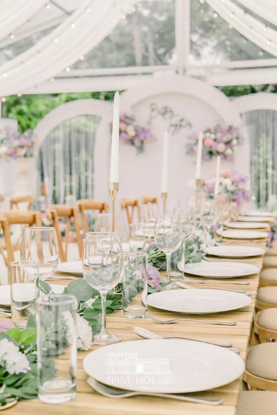 Long table set for a wedding reception, with flowers, candles, and white arches in the background.