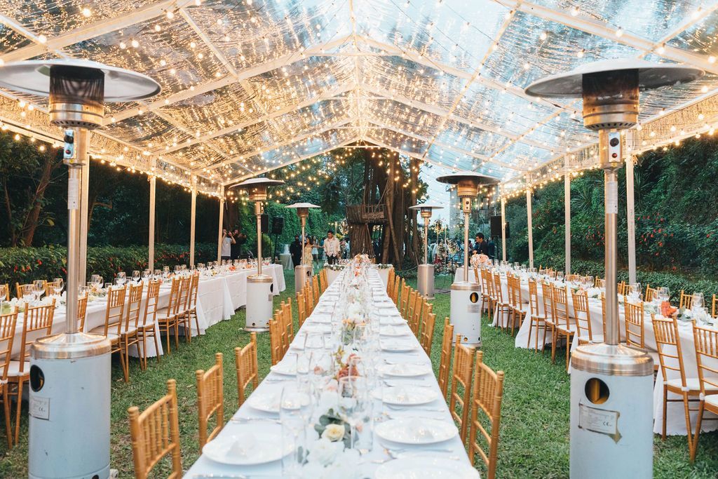 Long tables set for a celebration under a clear tent lit with string lights. Outdoor setting with heaters.